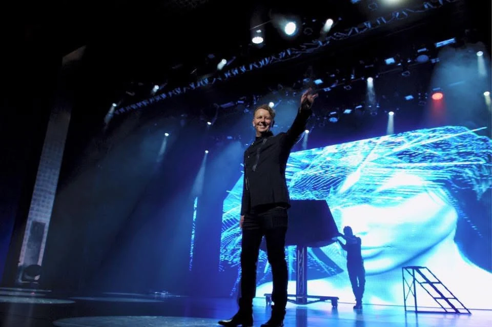 John Taylor acknowledging the audience on a theatre stage beneath blue lights