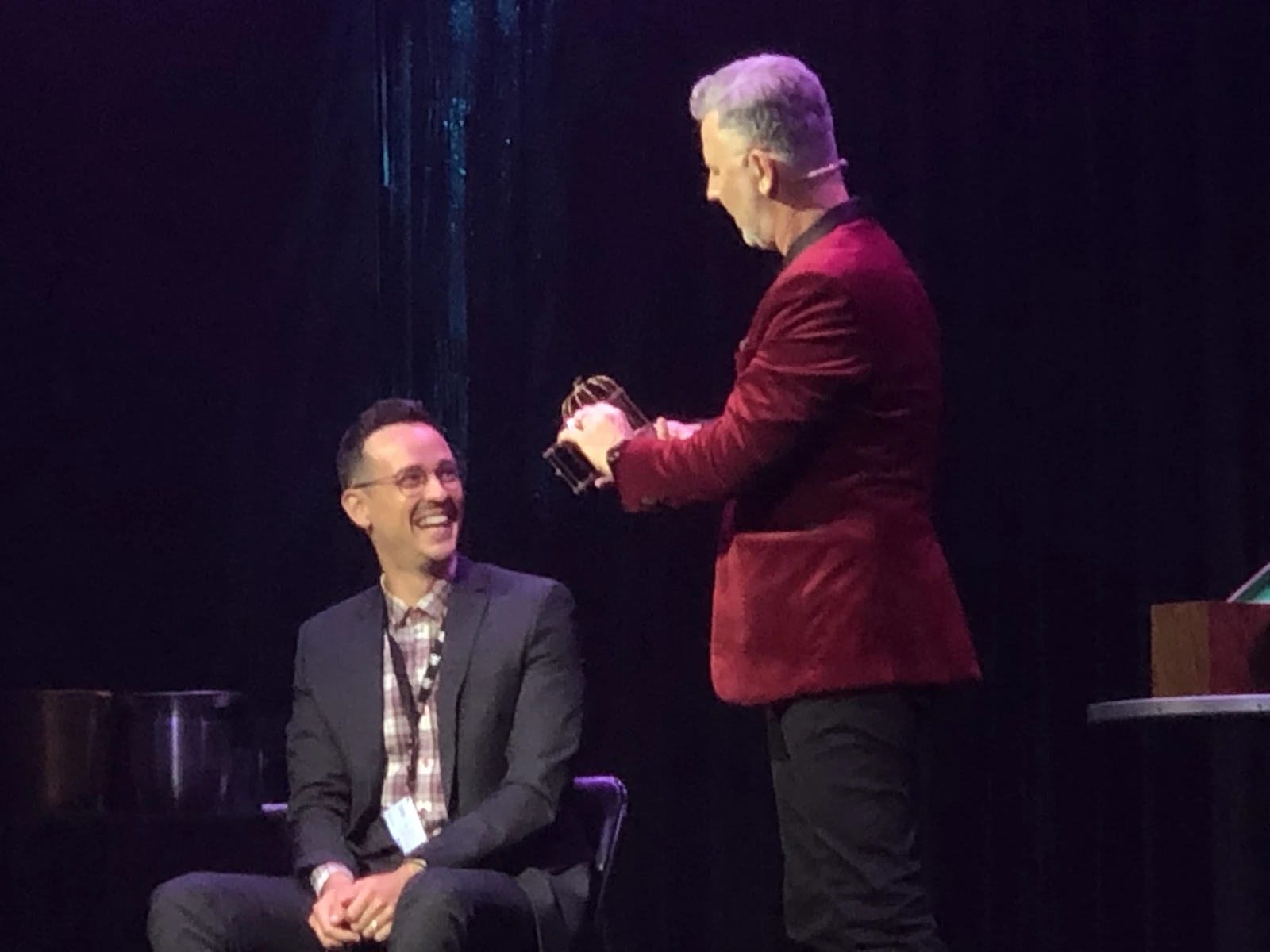John Taylor in a red jacket sharing a laugh with an audience volunteer on stage
